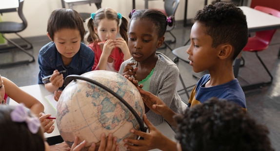 Kinder verschiedener Herkunft stehen in einem Klassenzimmer rund um einen Tischglobus.