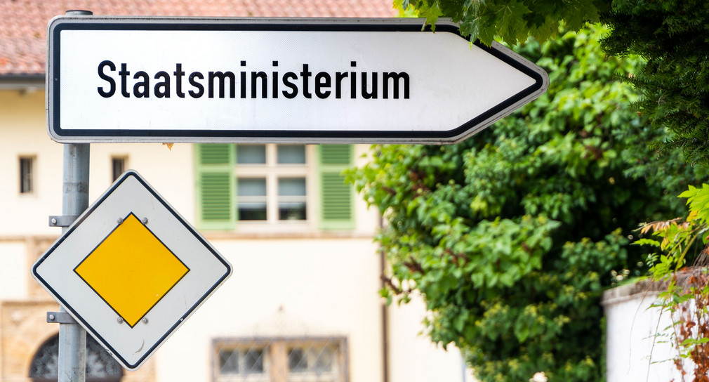 Ein Straßenschild mit der Aufschrift „Staatsministerium“ zeigt nach rechts. Darunter ist ein Schild „Vorfahrtsstraßse“ zu sehen.