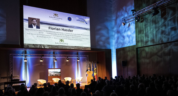 Blick in den Großen Saal der Landesvertretung. Die Gäste sitzen auf Stühlen. Auf dem Podium steht Staatssekretär Florian Hassler und hält seine Begrüßungsrede.