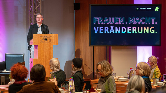 Staatssekretär Rudi Hoogvliet steht auf dem Podium hinter dem Rednerpult und begrüßt die Anwesenden