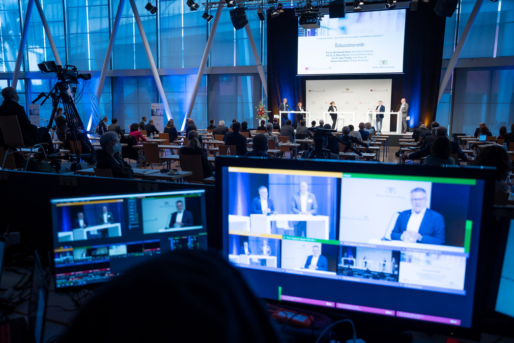 Im Vordergrund ein PC-Monitor, auf dem die Teilnehmenden einer Diskussionsrunde im Hintergrund auf der dritten Jahresveranstaltung des Forumgs Gesundheitsstandort Baden-Württemberg zu sehen sind.