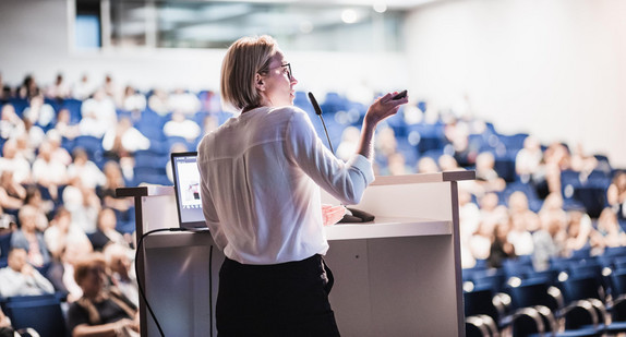Female speaker giving a talk on corporate business conference. Unrecognizable people in audience at conference hall. Business and Entrepreneurship event.