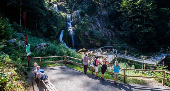Touristen stehen an einem Geländer und auf einer Aussichtsplattform und blicken auf eine Stufe der Triberger Wasserfälle, über die Wasser fließt.