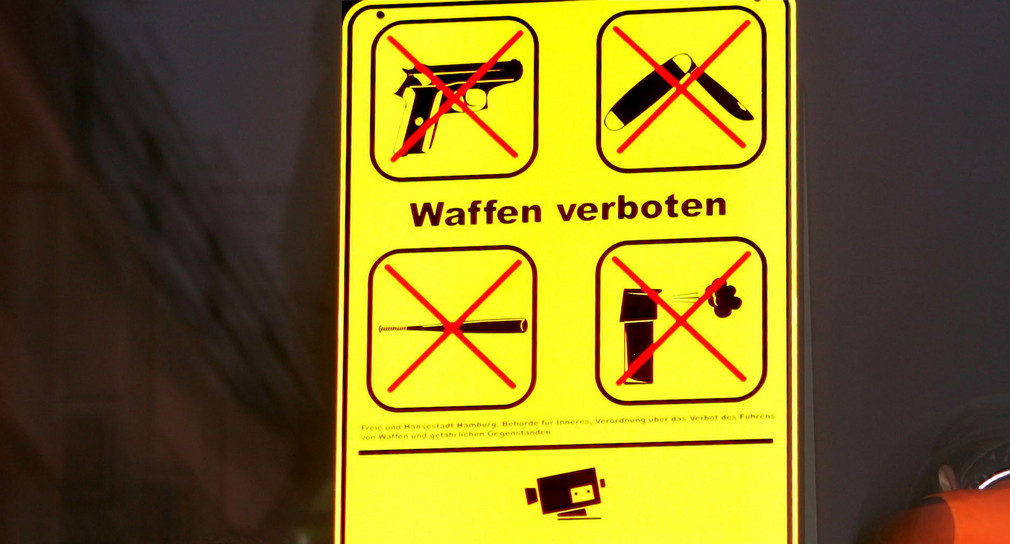 Vor nächtlichem Himmel ist ein gelbes Schild mit der Aufschrift „Waffen verboten“ zu sehen. Zudem befinden sich auf dem Schild durchgestrichene Piktogramme von einer Schusswaffe, einem Messer, einem Schlagstock und einer Reizgasdose.