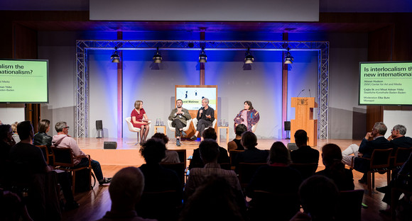 Blick in den großen Saal und auf das Podium mit Alistair Hudson (ZKM | Zentrum für Kunst und Medien Karlsruhe),  Çağla Ilk und Misal Adnan Yıldız (Staatliche Kunsthalle Baden-Baden) und Elke Buhr (Monopol). Der Saal ist dunkel, an den Seiten sind Lichstrahler eingesetzt die ein farbiges Muster an die Wand werfen. Rechts und links von der Bühne stehen zwei Plasmabildschirme mit dem Titel der Veranstaltung. Im Vordergrund sieht man schemenhaft die Köpfe des Publikums.