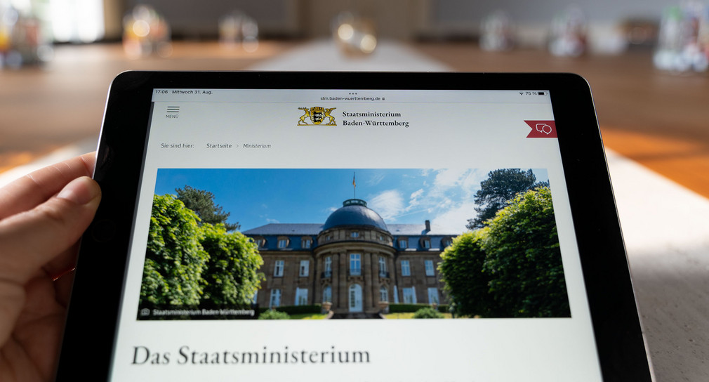 Eine Person sitzt im Kabinettssaal der Villa Reitzenstein und hält ein Tablett-Computer in der Hand. Auf dem Tablett-Computer ist die Webseite des Staatsministeriums zu sehen.