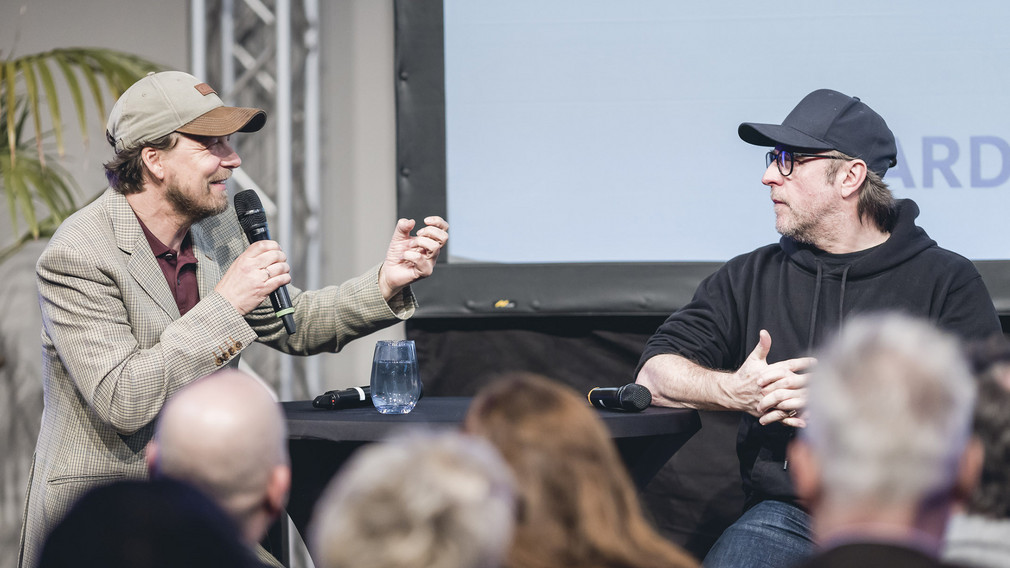 Regisseur Lars Jessen und Schauspieler Bjarne Mädel sitzen an Stehtischen auf Barhocker, Lars Jessen hält ein Mikrofon in der Hand