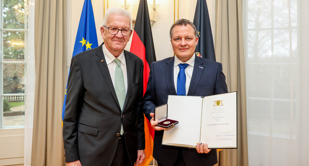 Ministerpräsident Winfried Kretschmann (links) und Thomas Speidel (rechts) mit der Staufermedaille in Gold und der Urkunde. Im Hintergrund stehen Fahnen.