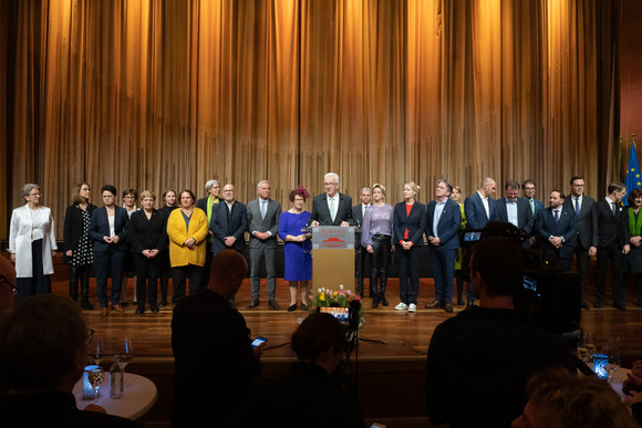 Ministerpräsident Winfried Kretschmann (Mitte) steht mit seiner Ehefrau Gerlinde und den Mitgliedern der Landesregierung auf der Bühne und spricht zu den Gästen.