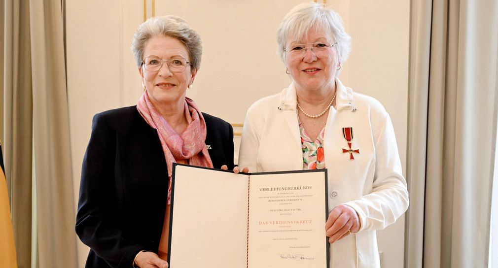 Zwei ältere Frauen, beide halten eine Mappe mit einer Urkunde, die andere trägt eine Auszeichnung am Revers.