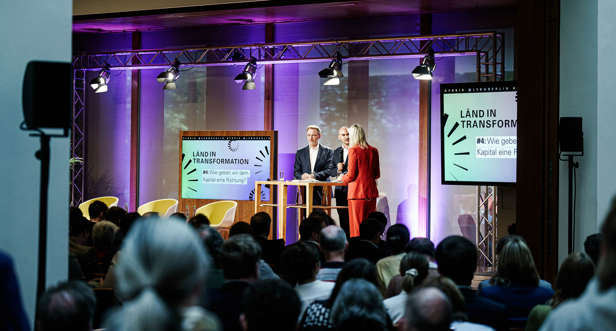 Seitlicher Blick in den Großen Saal über die Köpfe der Gästinnen und Gäste hinweg auf die Bühne, auf der Finanzminister Dr. Danyal Bayaz, Bundesfinanzminister Christian Lindner und die Moderatorin Sonja Álvarez Sobreviela zu sehen sind