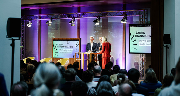 Seitlicher Blick in den Großen Saal über die Köpfe der Gästinnen und Gäste hinweg auf die Bühne, auf der Finanzminister Dr. Danyal Bayaz, Bundesfinanzminister Christian Lindner und die Moderatorin Sonja Álvarez Sobreviela zu sehen sind