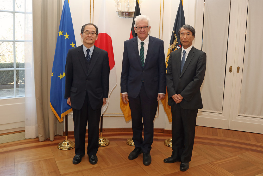 Der japanische Botschafter Hidenao Yanagi (links), Ministerpräsident Winfried Kretschmann (Mitte) und der japanische Generalkonsul Nobutaka Maekawa (rechts) stehen in der Villa Reitzenstein vor Fahnen.