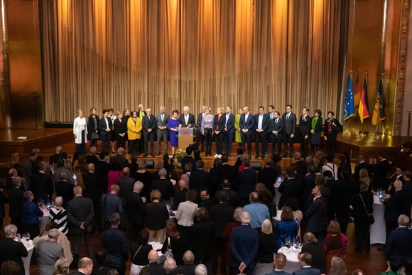 Ministerpräsident Winfried Kretschmann (Mitte) steht mit seiner Ehefrau Gerlinde und den Mitgliedern der Landesregierung auf der Bühne und spricht zu den Gästen.