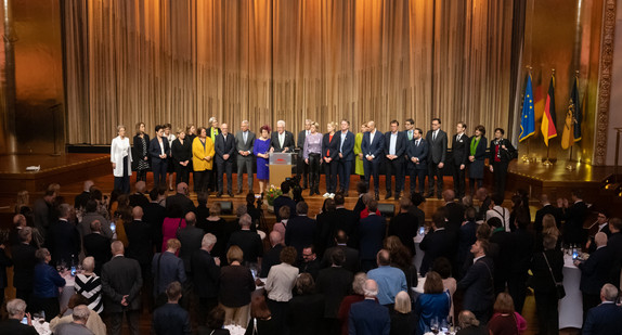 Ministerpräsident Winfried Kretschmann (Mitte) steht mit seiner Ehefrau Gerlinde und den Mitgliedern der Landesregierung auf der Bühne und spricht zu den Gästen.