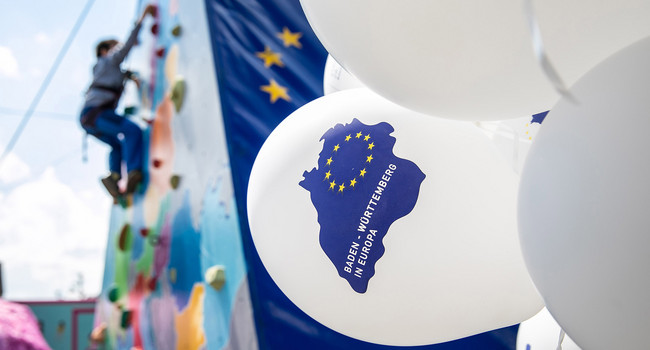 Einige weiße Luftballons mit der Aufschrift „Baden-Württemberg in Europa“ sind im Vordergrund zu sehen, im Hintergrund sind die EU-Flagge und eine Person an einer Kletterwand zu erkennen.