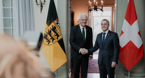 Ministerpräsident Winfried Kretschmann und Bundesrat Ignazio Cassis schütteln sich die Hand, flankiert von den Flaggen Baden-Württembergs und der Schweiz.