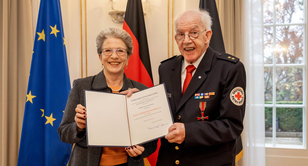 Auszeichnung Walter Adler mit Bundesverdienstkreuz geehrt Staatsrätin Barbara Bosch (links) und Walter Adler (rechts) stehen vor Fahnen und zeigen die Verleihungsurkunde.