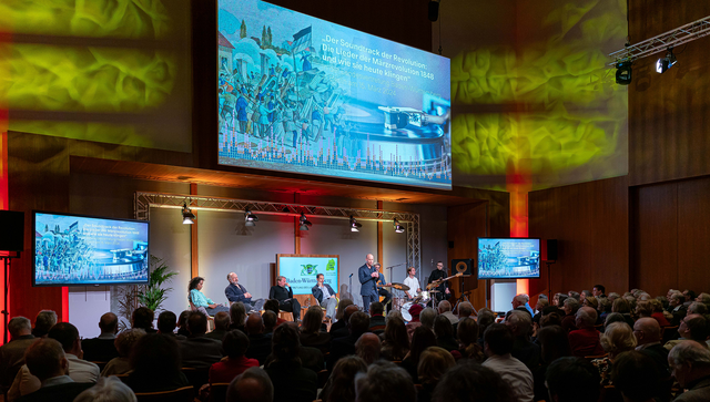 Blick in den Großen Saal der Landesvertretung mit dem Publikum, den Panelteilnehmerinnen und -teilnehmern und dem großen Transparent über der Bühne, auf dem der Titel der Veranstaltung und eine Szene der Märzrevolution und ein Schallplattenspieler zu sehen sind
