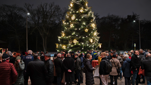 In der Mitte steht ein erleuchteter Weihnachtsbaum. Drumherum sind Menschen die sich unterhalten und gemeinsam Weihnachtsgeb&auml;ck essen.