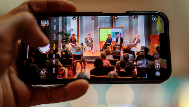 Eine Hand macht mit einem mobilen Endger&auml;t eine Aufnahme des Podiums.