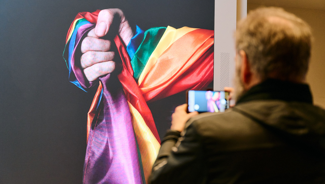 Einladungsbild der Veranstaltung auf der LED-Wand im Foyer der Landesvertretung. Ein Mann fotografiert die Hand, die eine Regenbogenfahne umschlungen hält